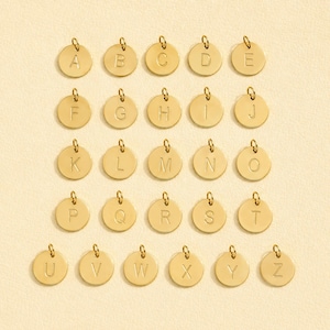 May include: A collection of twenty-six gold-toned circular charms, each featuring a single engraved uppercase letter from A to Z. Every charm includes a small jump ring at the top for attachment, arranged in a grid pattern on a light surface.