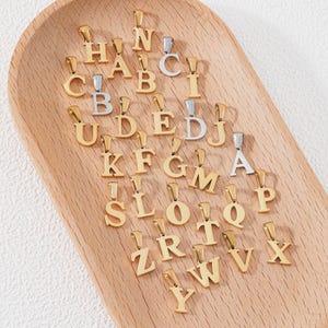 Könnte beinhalten: Eine Sammlung gold- und silberfarbener Alphabetanhänger, jeweils mit einer kleinen Schlaufe für eine Kette. Die Anhänger sind auf einem hellbraunen Holztablett angeordnet und zeigen Buchstaben von A bis Z.