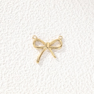 May include: A gold-toned metal charm shaped like a bow with a smooth, polished finish. The bow features two symmetrical loops and two dangling ribbon ends, with a small circular jump ring attached to each of the two top corners for connection.