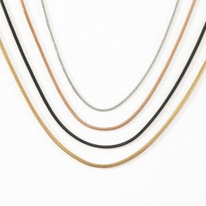 Può includere: Un set di quattro collane a catena snake disposte a cascata. La collezione comprende una catena color argento, una color oro rosa, una nera e una color oro, ognuna caratterizzata da un design a maglie lisce e flessibili.