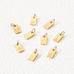 Pode incluir: Um conjunto de nove pequenos pingentes retangulares de metal dourado, cada um com uma letra gravada. As letras visíveis nos pingentes são U, F, S, O, L, A, B, J e E. Cada pingente inclui um pequeno anel circular na parte superior.