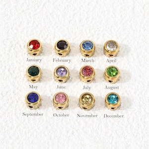 May include: Twelve gold-toned metal beads, each set with a different coloured gemstone representing a month of the year. The beads are arranged in a grid, with the corresponding month name printed in black text below each individual bead.