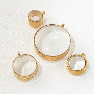May include: Four circular pendants of varying sizes, each featuring a clear, domed glass front encased in a polished gold-tone metal frame. Each pendant includes a small, integrated metal loop at the top for attachment to a chain or cord.