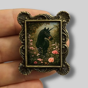 Op de afbeelding: Een kleine, rechthoekige broche met een gedetailleerde, antiek goudkleurige lijst. De ingelijste afbeelding toont een zwart eenhoorn in een veld met roze rozen. De eenhoorn heeft een lange, spiraalvormige hoorn en staat tegen een achtergrond van weelderig groen.