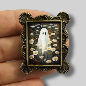 Può includere: Piccolo dipinto incorniciato raffigurante un fantasma bianco in un campo di margherite. La cornice è ornata, in stile bronzo antico. L'insieme evoca un tema stravagante e leggermente spettrale.
