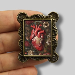 May include: A bronze-colored framed pin featuring a detailed anatomical heart illustration. The heart is red with visible veins, set against a floral background of red and white flowers. The frame has an ornate, scalloped edge.