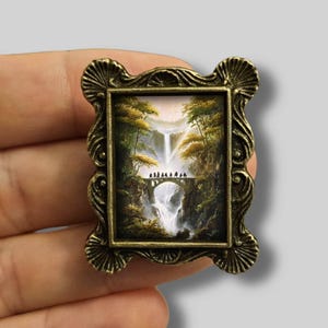 Op de afbeelding: Een miniatuur schilderij in een lijst dat een watervalscène met een brug en figuren afbeeldt. De lijst is versierd, met een bronzen afwerking. Het kunstwerk toont een waterval, bomen en een brug met kleine figuren, waardoor een gedetailleerd landschap ontstaat.
