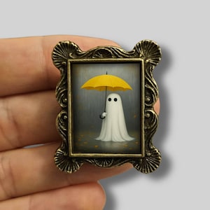 May include: A framed miniature painting of a white ghost holding a yellow umbrella in the rain. The frame is ornate with a bronze finish. The artwork has a whimsical, spooky theme.