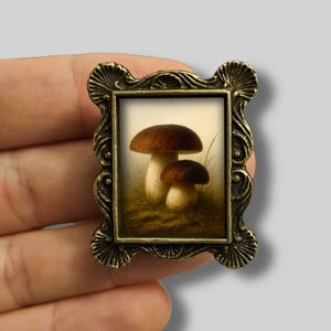 Może przedstawiać: Miniaturowe, oprawione dzieło sztuki przedstawiające obraz dwóch grzybów z brązowymi kapeluszami i białymi łodygami. Rama jest ozdobna z brązowym wykończeniem. Dzieło sztuki jest trzymane w dłoni.