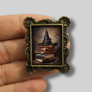 Puede incluir: Una pequeña pintura enmarcada que representa una pila de libros coronada por un sombrero de bruja negro. El marco es ornamentado, con un acabado bronce. La obra de arte tiene un tema oscuro y misterioso, centrado en la fantasía y la magia.