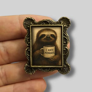 Op de afbeelding: Kleine, rechthoekige broche met een gedetailleerde, antiek goudkleurige lijst. De afbeelding toont een luiaard die een mok vasthoudt met de tekst "I HATE MONDAYS". De luiaard is bruin en de mok is wit.