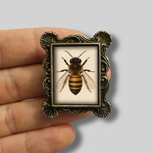 May include: A small, ornate, bronze-coloured frame featuring a detailed bee illustration. The bee has black and yellow stripes, wings, and antennae. The frame has a decorative, scalloped edge.