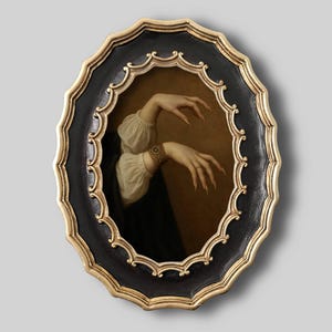 Puede incluir: Arte mural ovalado que presenta una pintura vintage de manos con uñas largas y puntiagudas. La obra de arte está enmarcada en un marco negro y dorado ornamentado, creando una estética gótica o vintage.