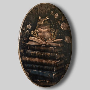 Puede incluir: Arte mural ovalado de madera con una ilustración detallada de un sapo sentado sobre una pila de libros. El sapo está rodeado de rosas y follaje, con una estética oscura y vintage. La obra de arte tiene un borde dorado.