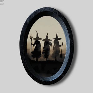 Könnte beinhalten: Ein ovaler schwarzer Holzrahmen mit einem Druck von drei silhouettierten Figuren mit spitzen Hüten. Die Figuren stehen vor einem nebligen, hellen Hintergrund, wobei eine einen Stab, eine einen kleinen Korb und eine einen Besen hält.