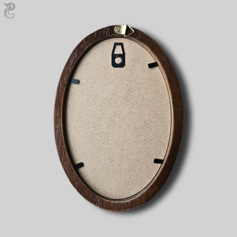 May include: An oval-shaped picture frame with a dark brown wood-grain finish. The back of the frame features a light tan fiberboard backing secured by four black metal clips, with a gold-toned metal triangular hanging hook attached at the top.