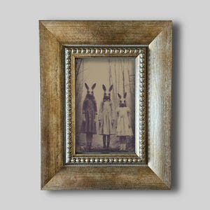 Könnte beinhalten: Gerahmter Vintage-Druck mit drei Figuren in Kleidern und Hasenmasken in einem Wald. Der Rahmen ist goldfarben mit einem perlenbesetzten Innenrand und einem strukturierten Außenrand.