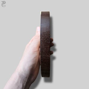 May include: A circular object with a dark brown, textured wooden surface viewed from the side. The item has a smooth, flat edge and a visible wood grain pattern throughout its exterior, showcasing a deep, rich brown color and a solid, sturdy construction.