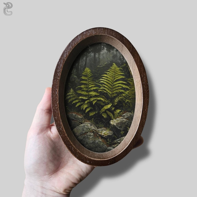 May include: An oval-shaped wooden frame containing a detailed painting of vibrant green ferns growing among grey rocks in a dark, dense forest setting. The frame has a smooth, dark brown finish and a simple, rounded edge design surrounding the artwork.