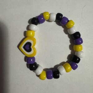 May include: A beaded bracelet featuring a yellow heart charm with a purple and black design. The bracelet is made of various colored beads, including yellow, white, black, and purple. The bracelet is a symbol of non-binary pride.