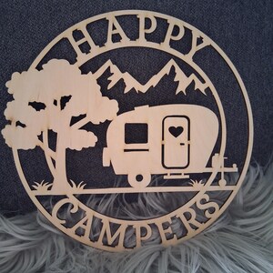 Könnte beinhalten: Ein kreisförmiges Holzschild mit den ausgeschnittenen Worten "HAPPY CAMPERS". Das Design umfasst einen Wohnwagen, Berge, einen Baum und ein Herz an der Wohnwagentür. Das Schild ist aus hellem Holz gefertigt.