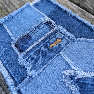 May include: A patchwork denim fabric with frayed edges. The fabric is made of different shades of blue denim and has a small embroidered design in the center.