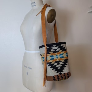 May include: A white, black, brown, blue, and yellow patterned tote bag with a long brown leather strap and fringe. The bag is hanging on a white dress form.