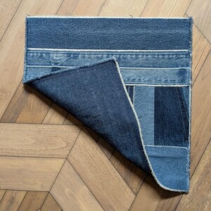 Pu&ograve; includere: Un tappeto rettangolare in denim realizzato con jeans riciclati. Il tappeto presenta varie tonalit&agrave; di denim blu, con cuciture visibili e un angolo ripiegato che rivela il lato inferiore. Il tappeto &egrave; posizionato su un pavimento in legno.