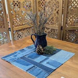 May include: A square patchwork denim fabric mat featuring various shades of blue, topped with a dark blue ceramic pitcher filled with dried lavender stems, a single brown pinecone, and a small sprig of green foliage on a wooden surface.