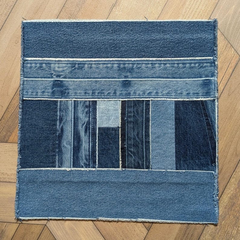 Pu&ograve; includere: Tessuto patchwork quadrato in denim. Il tessuto &egrave; realizzato con varie tonalit&agrave; di denim blu, disposte a strisce orizzontali e verticali. I bordi sono sfilacciati e il pezzo &egrave; appoggiato su una superficie di legno.