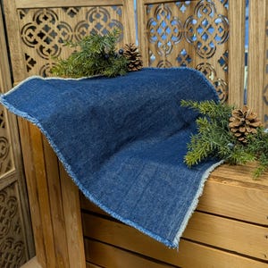 Pu&ograve; includere: Un sottopentola quadrato in denim blu indaco con bordo sfilacciato. Il sottopentola &egrave; esposto su una superficie di legno e decorato con rami di pino e pigne.