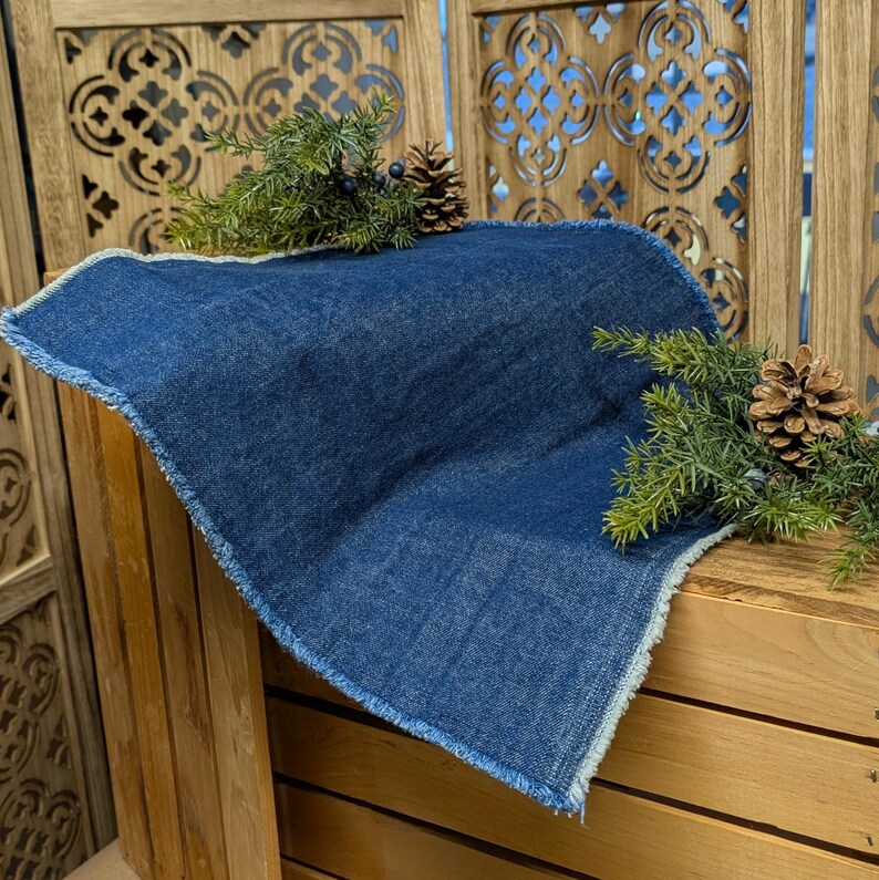 Pu&ograve; includere: Un sottopentola quadrato in denim blu indaco con bordo sfilacciato. Il sottopentola &egrave; esposto su una superficie di legno e decorato con rami di pino e pigne.