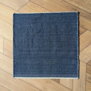 Pu&ograve; includere: Un tessuto quadrato di denim color indaco con un bordo sfilacciato. Il tessuto &egrave; di colore uniforme e sembra un piccolo tappeto o una tovaglietta. Lo sfondo &egrave; un pavimento in legno marrone chiaro.