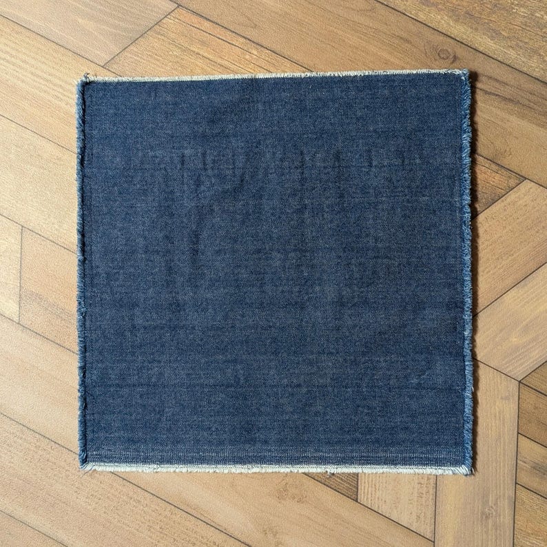 Pu&ograve; includere: Un tessuto quadrato di denim color indaco con un bordo sfilacciato. Il tessuto &egrave; di colore uniforme e sembra un piccolo tappeto o una tovaglietta. Lo sfondo &egrave; un pavimento in legno marrone chiaro.