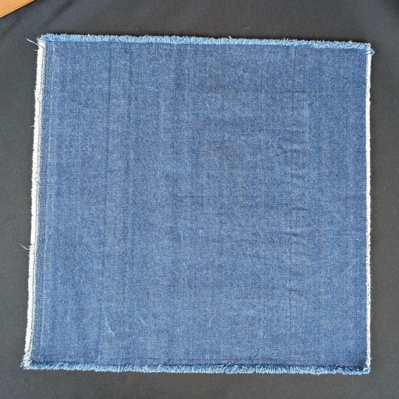 Pu&ograve; includere: Un campione di tessuto quadrato blu denim con un bordo sfilacciato. Il tessuto ha un aspetto strutturato e un colore blu medio. I bordi sono rifiniti con un bordo bianco e un bordo sfilacciato.