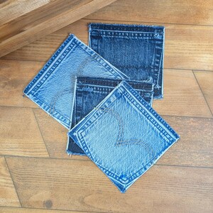 May include: A set of three square coasters crafted from denim fabric, featuring varying shades of blue from light to dark. Each coaster displays visible stitching patterns and frayed edges, mimicking the appearance of classic jean pockets.