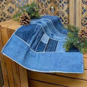 May include: A denim table runner with a patchwork design. The runner features various shades of blue denim, with a lighter blue border and a frayed edge. The runner is displayed on a wooden crate, decorated with pinecones and greenery.
