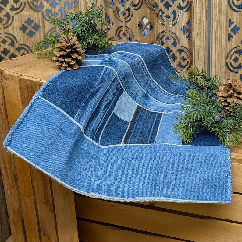 Pu&ograve; includere: Una tovaglia in denim con un design patchwork. La tovaglia presenta varie tonalit&agrave; di denim blu, con un bordo blu pi&ugrave; chiaro e un bordo sfilacciato. La tovaglia &egrave; esposta su una cassa di legno, decorata con pigne e verde.