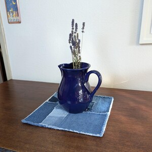 May include: A dark blue ceramic pitcher with a handle, filled with dried lavender flowers, sits on a blue denim patchwork coaster.