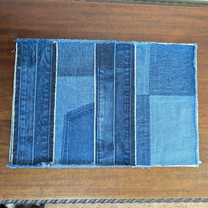 May include: A blue denim patchwork placemat with a frayed edge. The placemat is made from different pieces of denim sewn together.