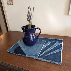 May include: A blue ceramic pitcher with dried lavender flowers sits on a blue denim placemat with frayed edges.