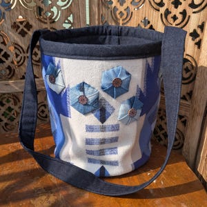 May include: A handmade bucket bag with a white and blue patterned design. The bag features denim accents, including the strap and decorative flower appliqués with wooden button centers. The bag is designed with a denim trim and a denim interior.