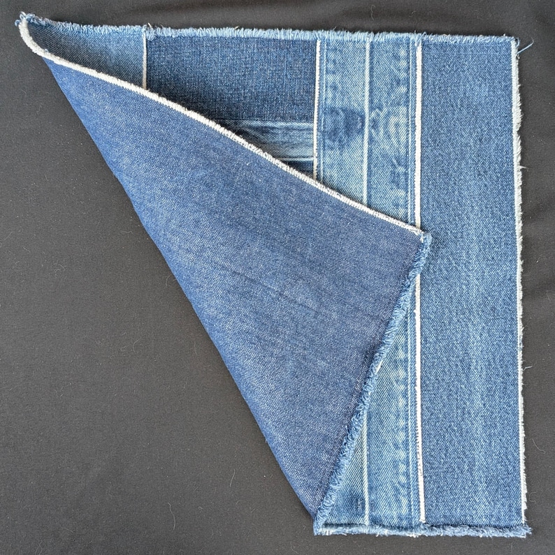 Pu&ograve; includere: Un pezzo quadrato di tessuto denim con un angolo piegato, che mostra varie tonalit&agrave; di denim blu. Il design incorpora strisce verticali con cuciture bianche e una finitura a bordo grezzo. L'angolo piegato rivela l'interno del tessuto.