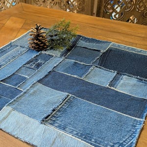 May include: A square placemat constructed from various shades of blue denim fabric patches with frayed edges. The patchwork design features rectangular and square pieces of denim stitched together. A brown pinecone and green sprig rest on top.