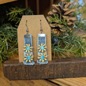 May include: Denim earrings with embroidered yellow and blue flowers. The earrings are rectangular with silver-colored metal accents and hooks. They are displayed on a brown card.
