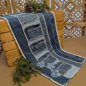 May include: A denim table runner with a patchwork design. The runner is made from various shades of blue denim, with frayed edges and decorative butterfly accents. A pinecone and greenery add a rustic touch.