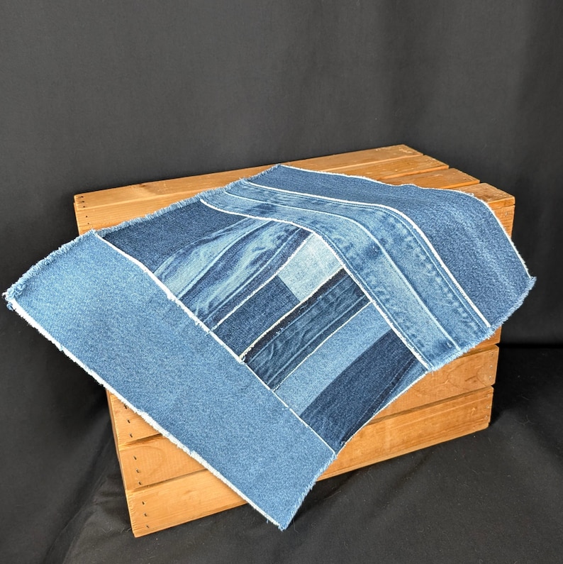 Pu&ograve; includere: Un articolo quadrato in patchwork di denim con varie tonalit&agrave; di blu. Il design patchwork presenta pezzi rettangolari e quadrati, con cuciture bianche. L'articolo &egrave; posto su una cassa di legno su sfondo nero.