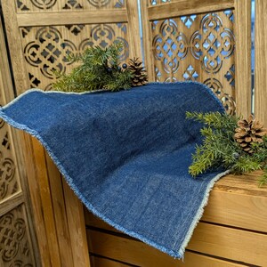 Pu&ograve; includere: Un sottopentola quadrato blu denim con un bordo sfilacciato. Il sottopentola &egrave; decorato con rametti di aghi di pino verdi, piccole bacche scure e pigne. Il sottopentola &egrave; appoggiato su una superficie di legno.