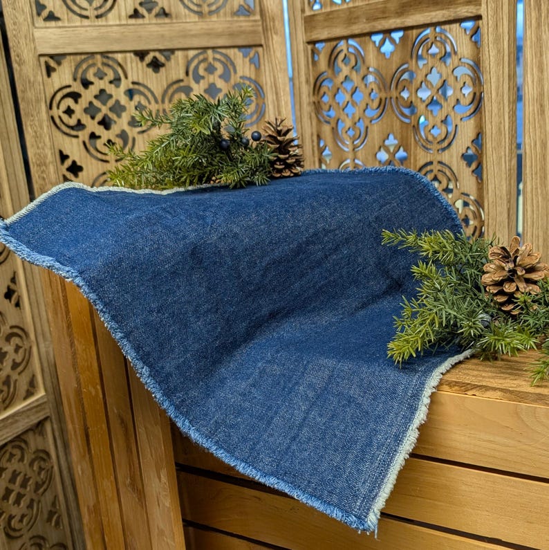 Pu&ograve; includere: Un sottopentola quadrato blu denim con un bordo sfilacciato. Il sottopentola &egrave; decorato con rametti di aghi di pino verdi, piccole bacche scure e pigne. Il sottopentola &egrave; appoggiato su una superficie di legno.
