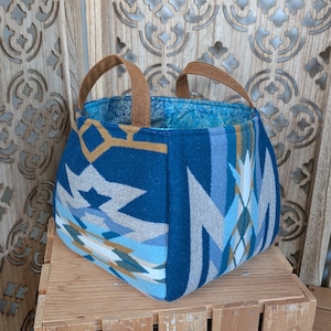 May include: A square-shaped tote bag with a Southwestern-inspired pattern in shades of blue, gray, and tan. The bag features brown leather handles and a patterned blue interior. The bag is sitting on a wooden surface.